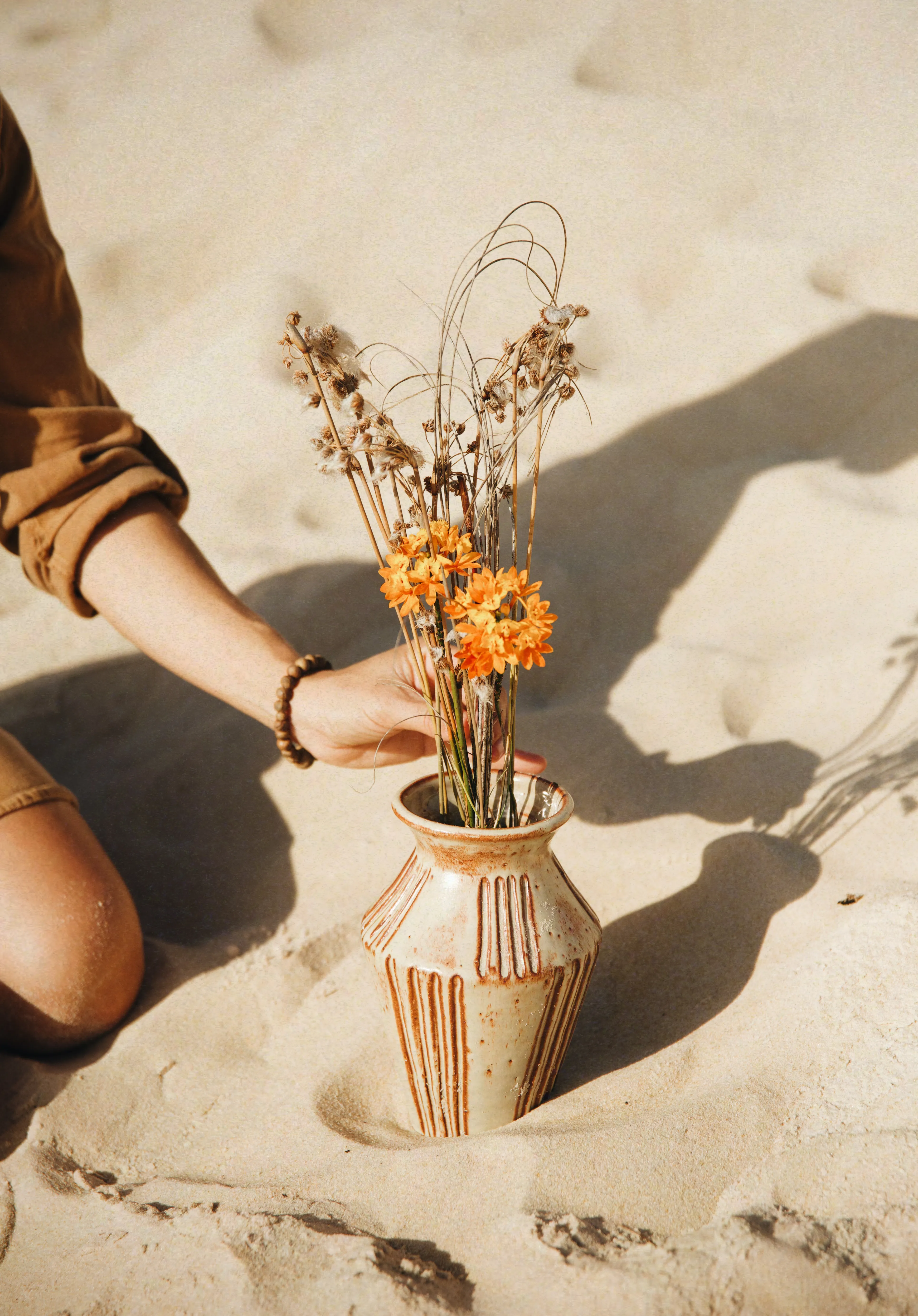 Mão arrumando flores laranja no vaso Dunas #03 assentado na areia, grafismos verticais incisos