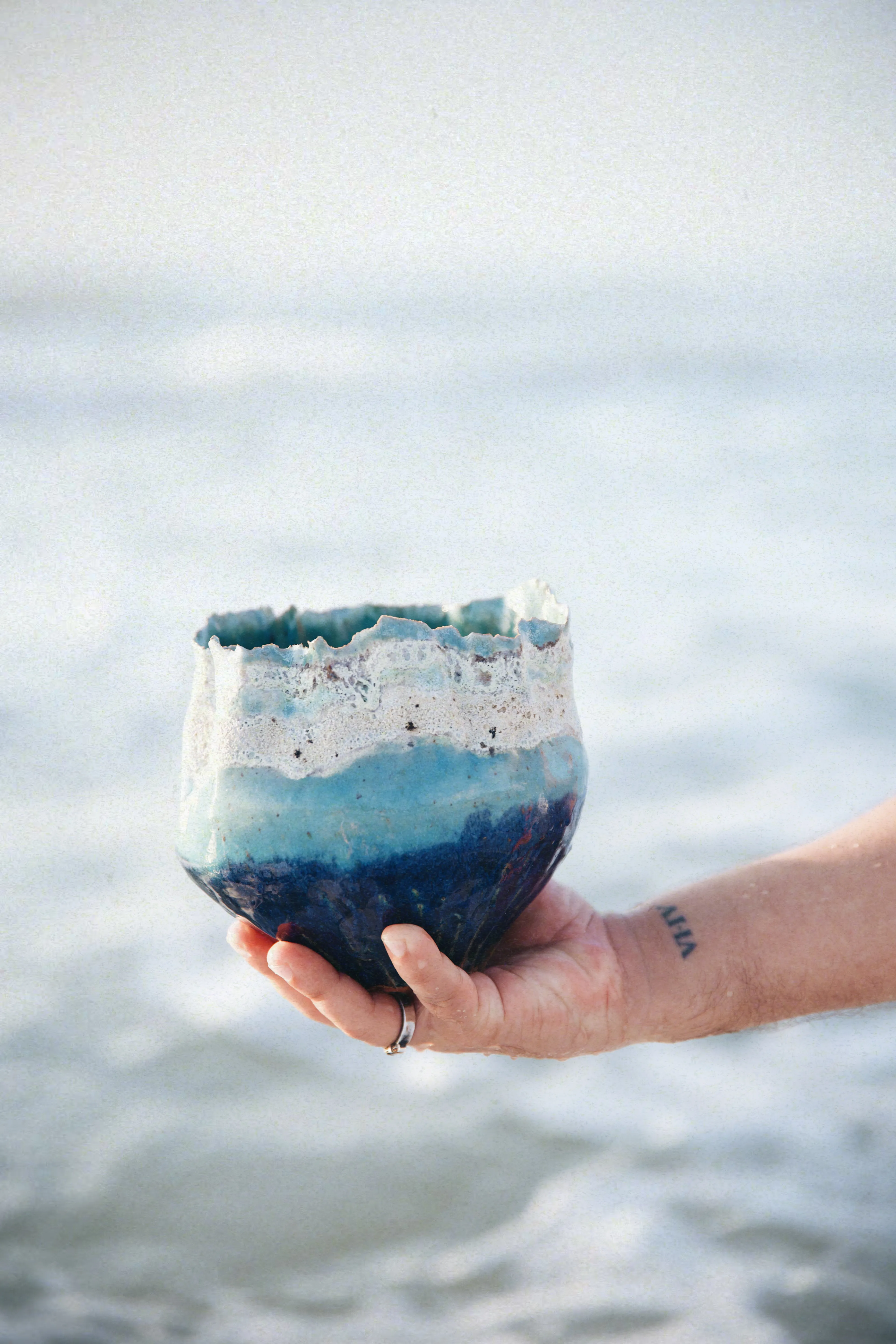 Tigela Mar #01 sustentada por uma mão diante do mar, com bordas vivas e esmalte em camadas de azul